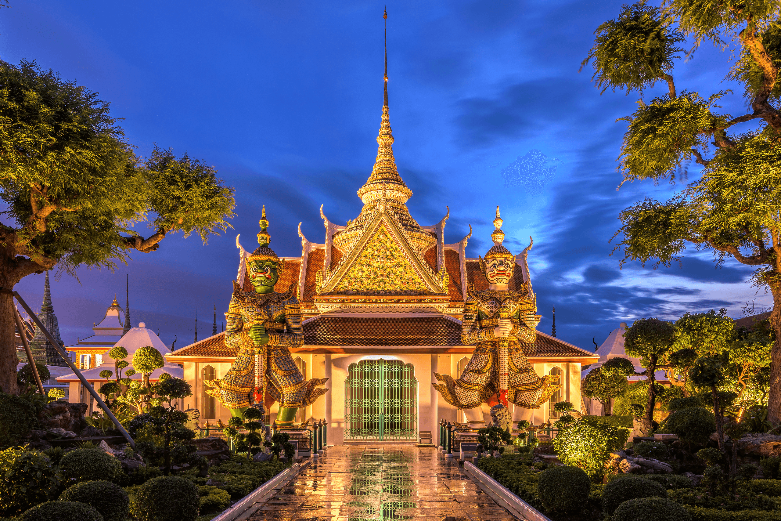 A pavilion with a gilded roof framed by two large guardian statues, lit at dusk.